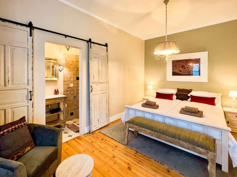 Ein Schlafzimmer mit einem Doppelbett, einem Sessel und einem angrenzenden Badezimmer, das durch eine Schiebetür aus Holz abgetrennt ist.