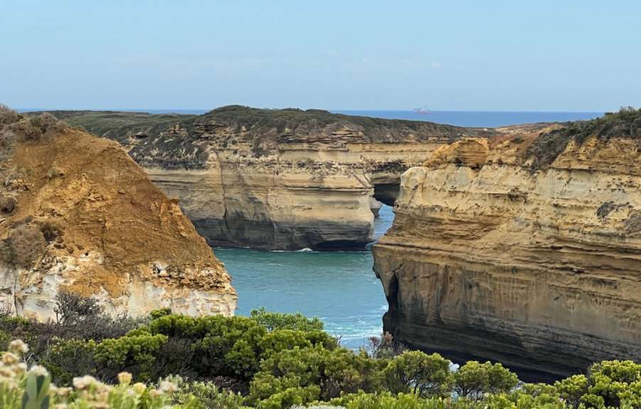 Ein Schiff fährt auf dem Meer hinter zerklüfteten Felsformationen und grüner Vegetation.