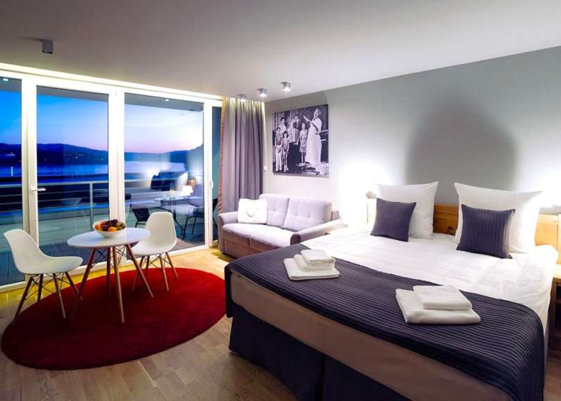 Ein geräumiges Hotelzimmer mit großem Fenster, das einen Blick auf das Wasser bietet, einem Bett, einer Couch und einem kleinen Tisch mit Stühlen.