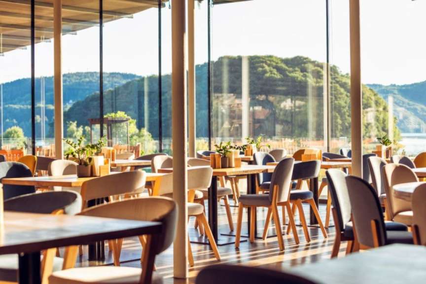 In einem hellen Restaurant mit großen Fenstern stehen mehrere Tische und Stühle, im Hintergrund ist eine grüne Hügellandschaft mit Wasser zu sehen.
