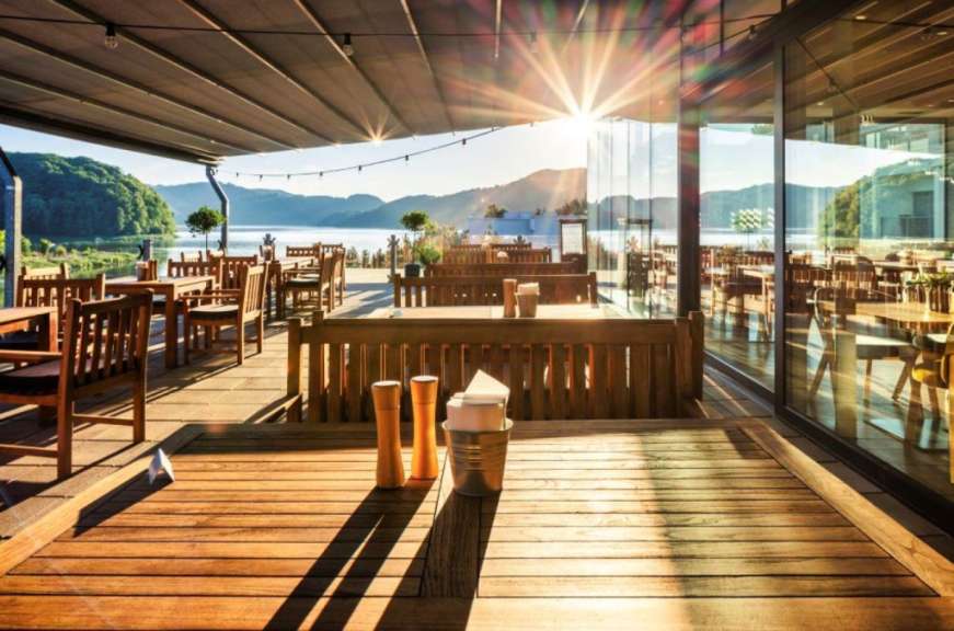 Eineische Restaurantterrasse mit Holztischen und Stühlen bietet einen Ausblick auf einen sonnigen See und bewaldete Hügel.