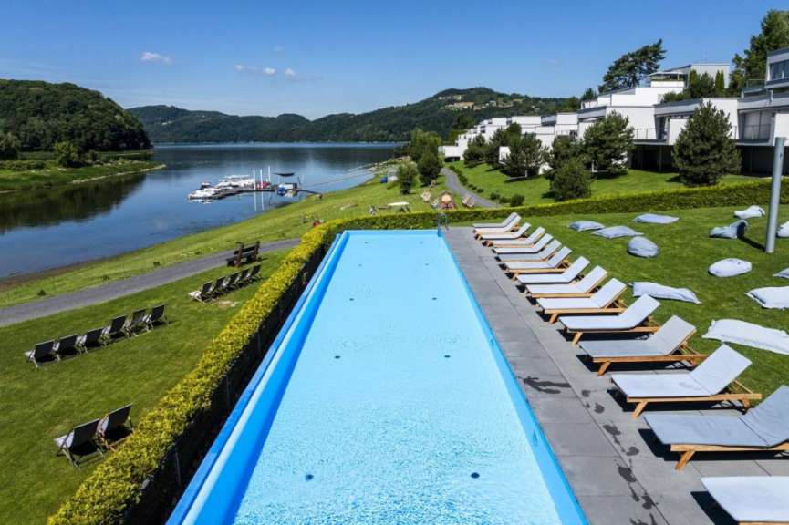 Ein langer, blauer Swimmingpool ist auf einem grünen Rasen mit Liegestühlen und Sitzsäcken angeordnet, mit einem See und bewaldeten Hügeln im Hintergrund.