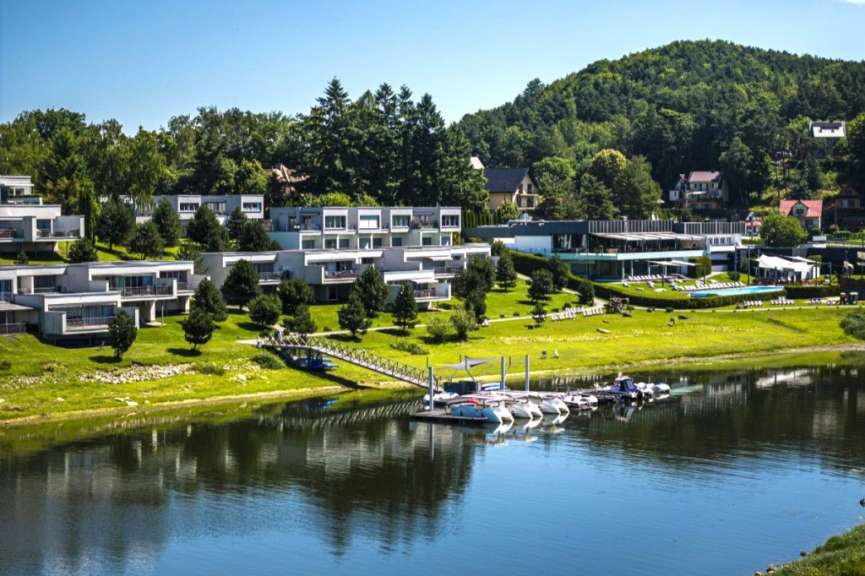 Ein modernes Hotel mit einer breiten Glasfassade und einem Pool liegt am Ufer eines ruhigen Sees, der von einer grünen Hügelkette und umliegenden Häusern umgeben ist.