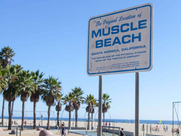 Ein blau-weißes Schild an einem Strand, das die ursprüngliche Lage des Muskelstrandes in Santa Monica, Kalifornien zeigt