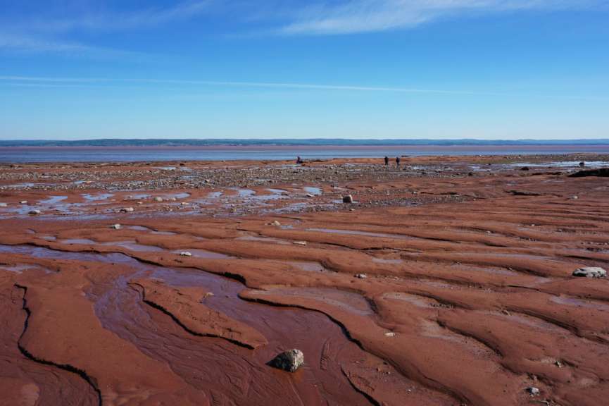 Ein breiter, von Wasserläufen durchzogener Sandstrand mit Felsen und einer entfernten Hügellandschaft unter blauem Himmel.