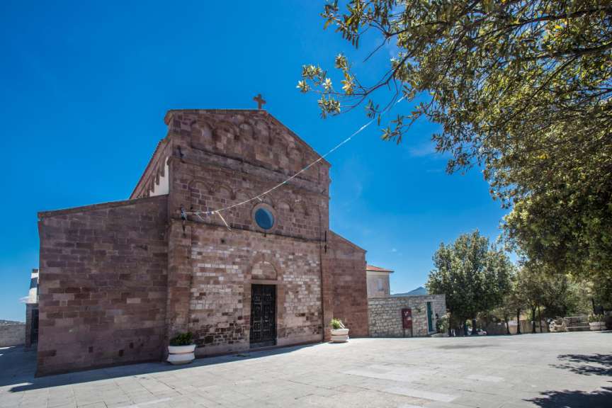 Ein kirchliches Gebäude aus Ziegelstein steht auf einem Platz im Freien unter einem blauen Himmel, mit Bäumen, die von rechts ins Bild ragen.