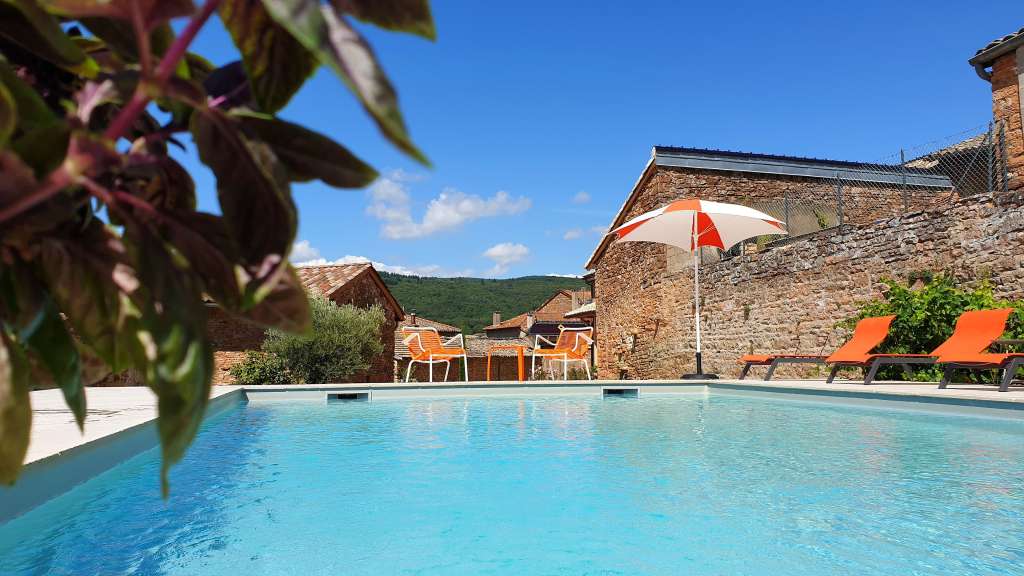 Ein heller, sonniger Tag mit einem türkisfarbenen Pool, umgeben von Steinbauten, gepunktet mit orangefarbenen Liegestühlen und einem Sonnenschirm.