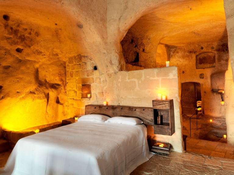 Ein Bett mit weißen Laken steht in einem beleuchteten Höhlenraum, der mit Steinmauern und Decken ausgekleidet ist.
