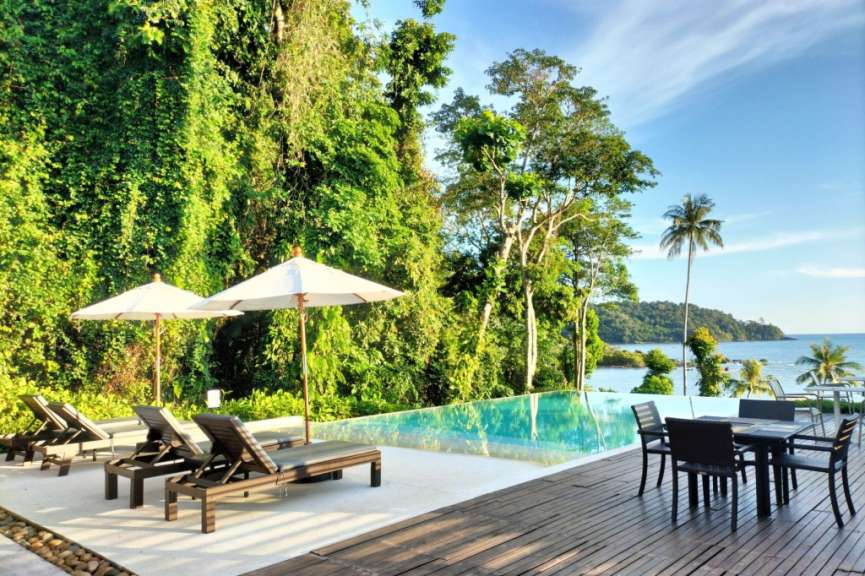 Ein Infinity-Pool mit Liegestühlen und Sonnenschirmen steht vor einer tropischen Dschungelwand mit Blick auf das Meer und eine Insel.