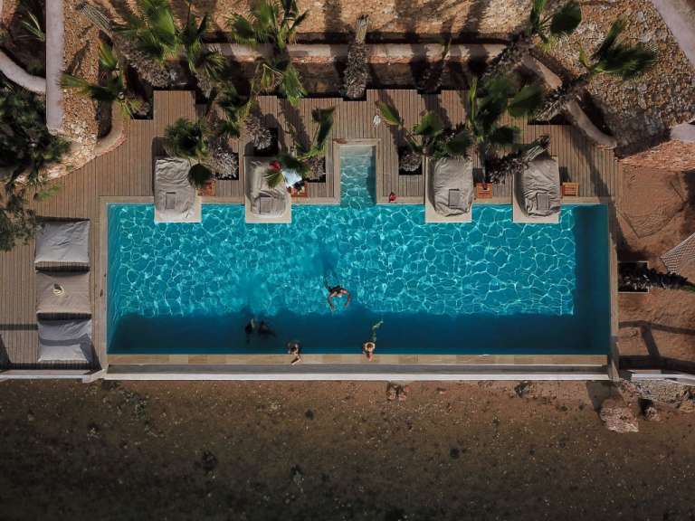 Eine Drohnenansicht zeigt ein Schwimmbad im Freien mit Menschen, die schwimmen, und Liegestühlen im Schatten von Palmen.