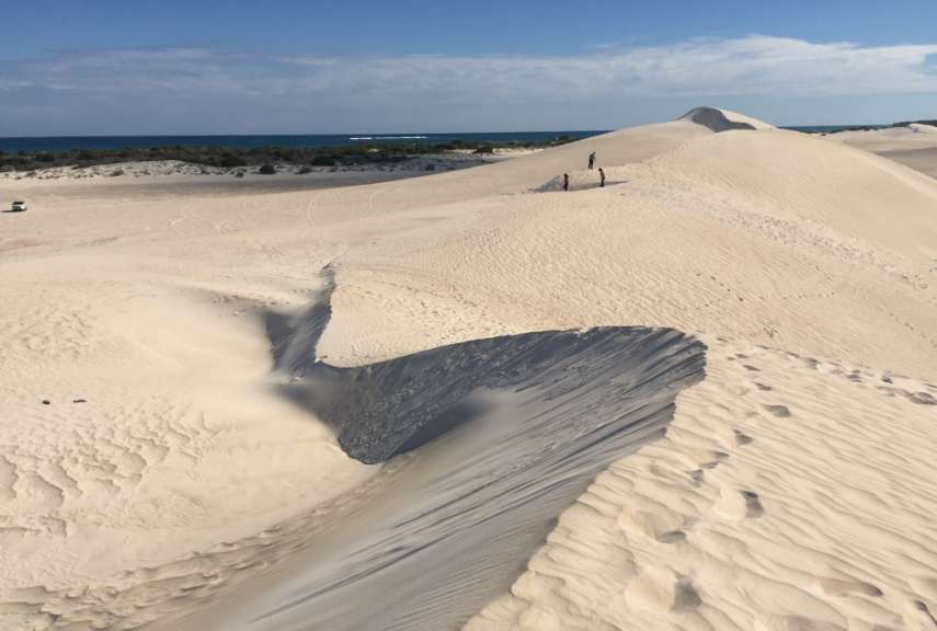 Drei Personen stehen auf einer Sanddüne am Meer, während ein Auto in der Ferne fährt.