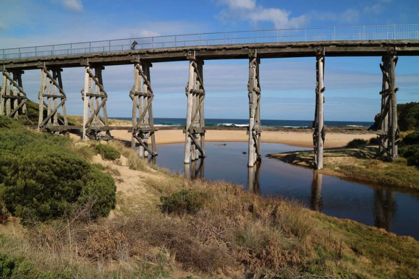 Ein hölzerner Viadukt überspannt einen Flusslauf am Meer unter einem blauen Himmel mit Wolken.