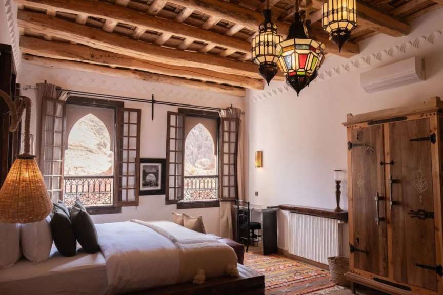 Ein Schlafzimmer im orientalischen Stil mit Holzbalken an der Decke, bunten Laternen und einem Bett mit Blick auf eine felsige Landschaft.
