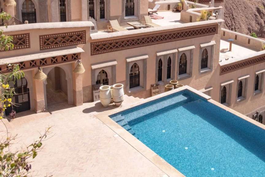 Ein moderner Pool mit türkisfarbenem Wasser und hellbraunen Fliesen ist von einem traditionellen Gebäude mit marokkanischer Architektur umgeben.