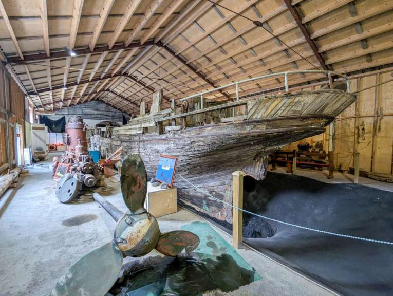 Ein alter Holzschiffskörper steht in einer Lagerhalle auf einem Betonboden, daneben liegen ein großes Propellerblatt und Maschinen.