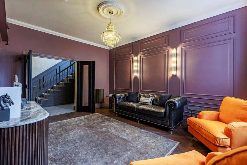In einem luxuriösen Wohnzimmer mit lila Wänden und Parkettboden stehen ein schwarzes Ledersofa und zwei orangefarbene Sessel vor einem gemusterten Teppich.