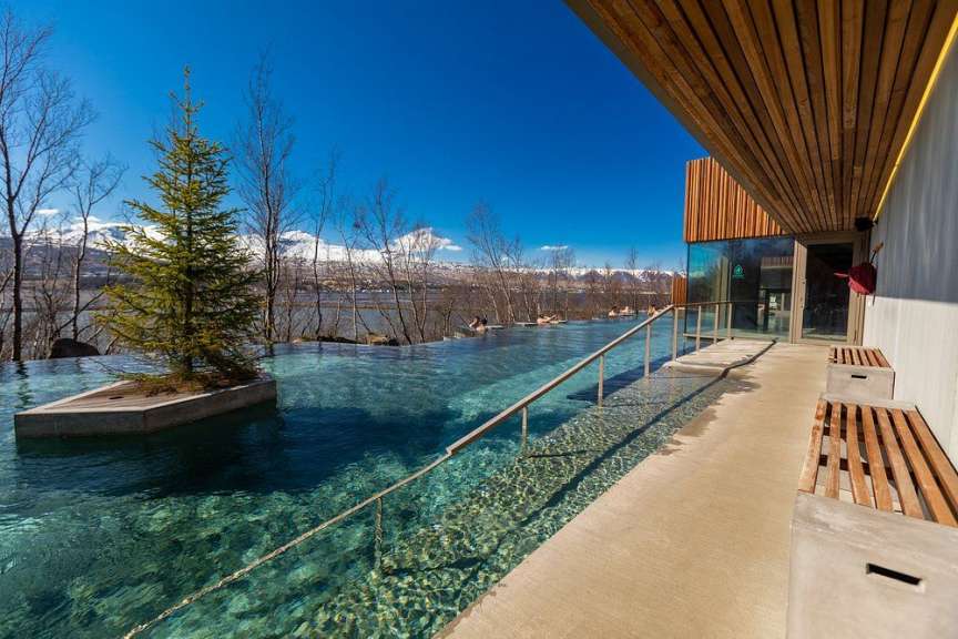 Ein Infinity-Pool mit Blick auf schneebedeckte Berge und einen See, daneben eine moderne Architektur mit Sitzgelegenheiten.