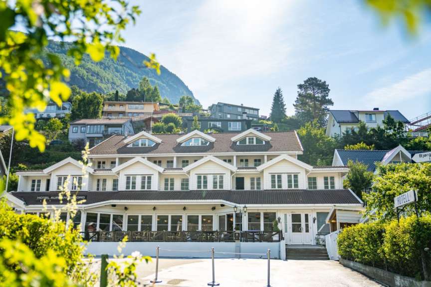Ein großes weißes Hotel mit vielen Fenstern und Balkonen ist auf einem Hügel mit Bäumen im Vordergrund eines blau bewölkten Himmels zu sehen.