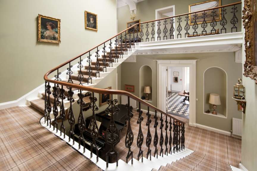 Eine geschwungene Treppe mit kariertem Teppich führt nach oben, flankiert von einer verzierten Balustrade und Wandgemälden.
