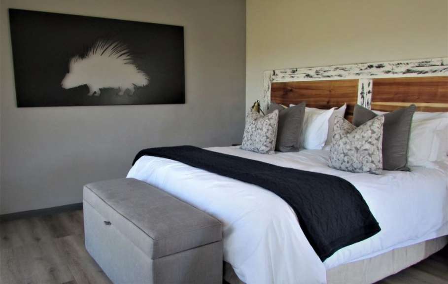 Ein Schlafzimmer mit einem Bett, das mit weißer Bettwäsche, mehreren Kissen und einer schwarzen Decke bedeckt ist, einem Nachttisch aus grauem Stoff, der vor dem Bett steht, und einem großen rechteckigen Bild an der Wand, das ein weißes Stachelschwein auf schwarzem Hintergrund zeigt.