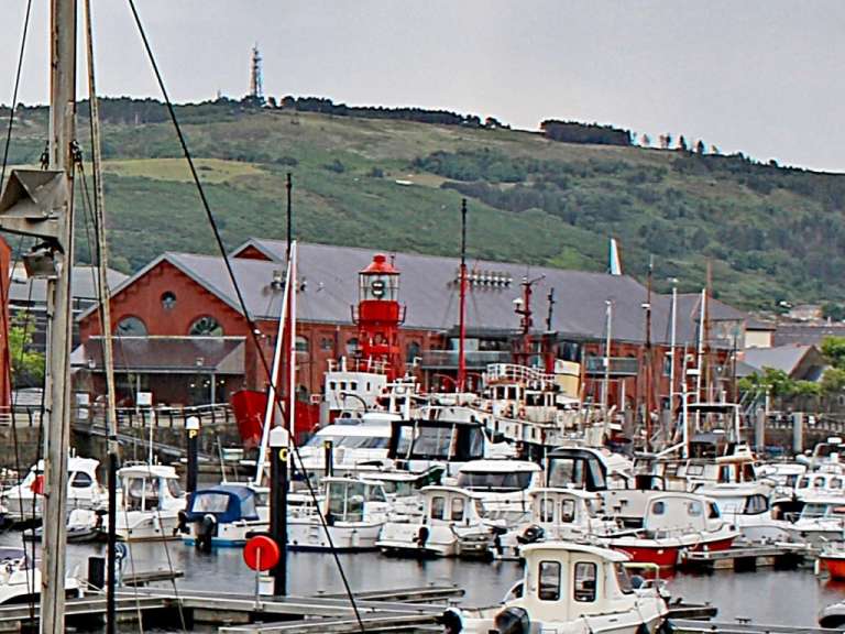 Ein Blick auf einen Yachthafen mit vielen Booten, einem roten Leuchtturm und einem Hügel im Hintergrund.