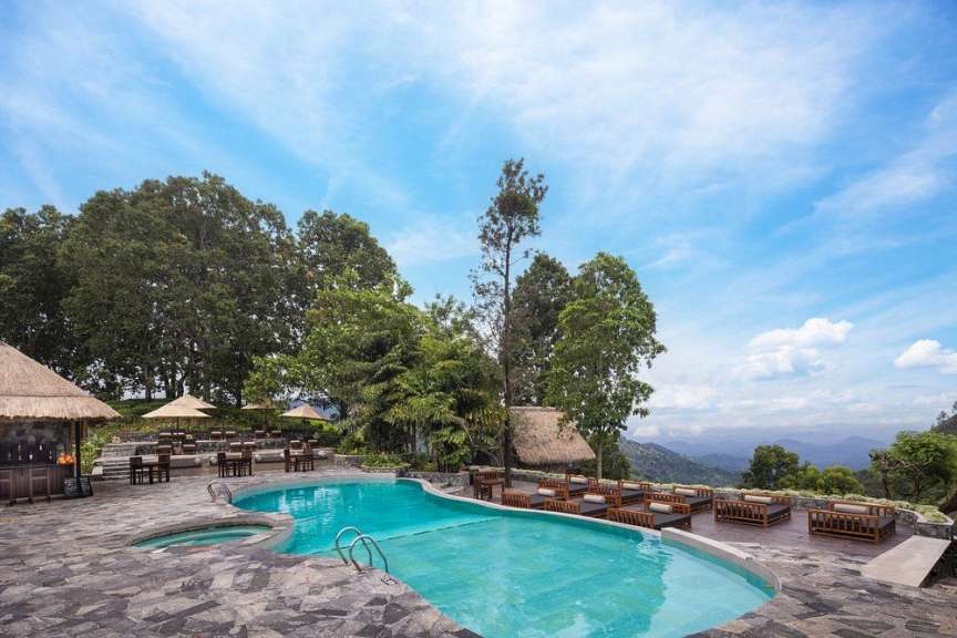 Ein großer Pool mit türkisfarbenem Wasser ist von einem steinernen Terrassenbereich umgeben, auf dem Liegestühle, Tische und Stühle aufgestellt sind, während im Hintergrund üppige Bäume und eine bergige Landschaft zu sehen sind.