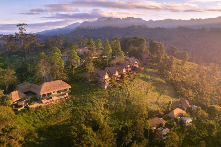 Eine Ansammlung von strohgedeckten Häusern und Gebäuden ist auf einem Hügel in einer bergigen Landschaft zu sehen, mit Bäumen und Terrassen, die die Hänge bedecken, und einem wolkenverhangenen Himmel im Hintergrund.