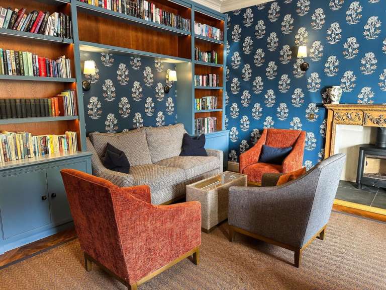 Eine gemütliche Leseecke mit Sofas, Sesseln, einem Kamin und Bücherregalen, die mit vielen Büchern gefüllt sind, schmückt dieses Bild.