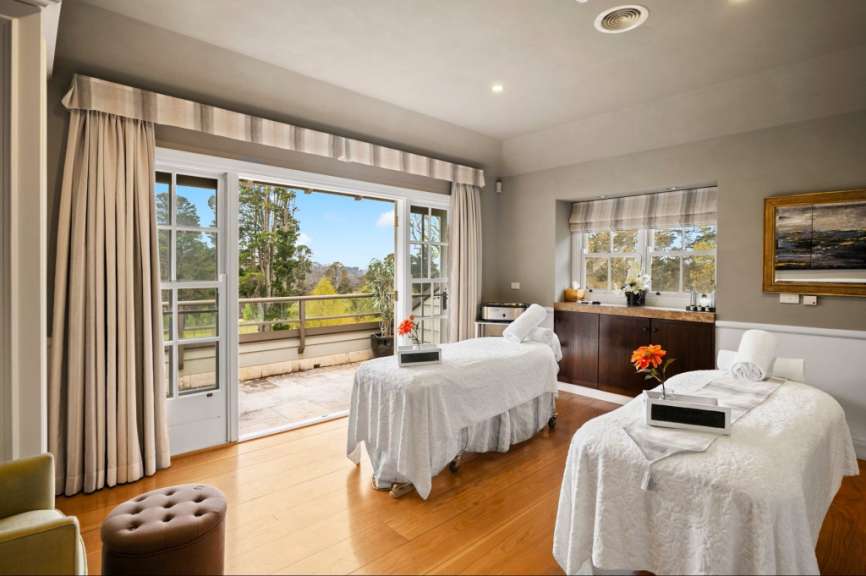 Das Bild zeigt ein luxuriöses Spa-Zimmer mit zwei Massageliegen, die mit weißen Laken bedeckt sind, und einem großen Schiebefenster mit Blick auf eine üppige Landschaft.