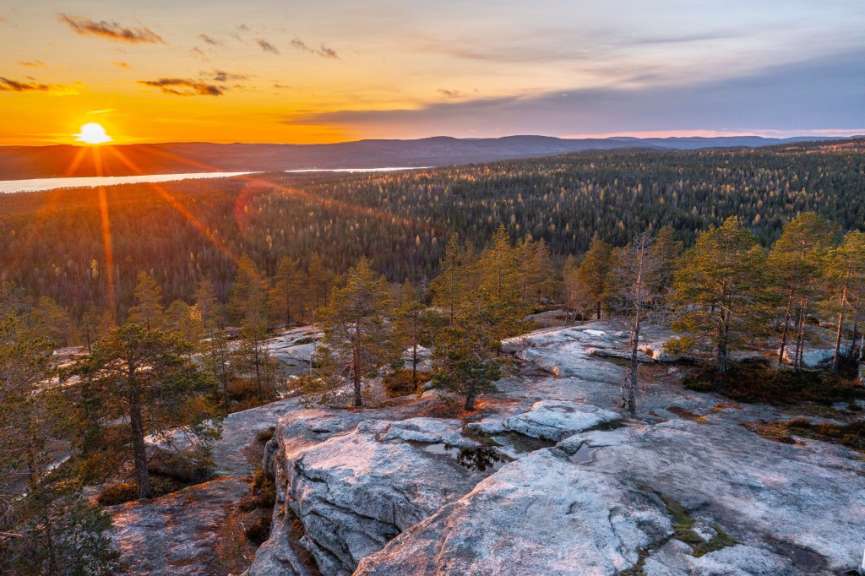 Ein Sonnenuntergang beleuchtet eine felsige Landschaft mit Kiefern und einem Wald im Hintergrund.