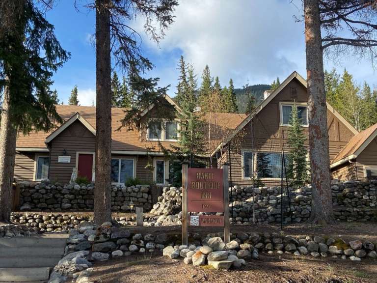 Ein braunes Holzhaus mit einem roten Eingang, umgeben von Bäumen und einem Steinfundament, wirbt mit einem Schild für das "Banff Boutique Inn".