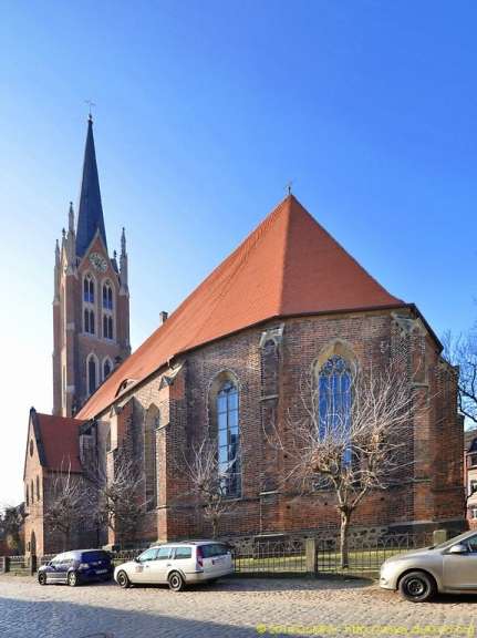 Eine hohe Backsteinkirche mit rotem Ziegeldach und einem spitz zulaufenden Turm steht unter einem blauen Himmel.