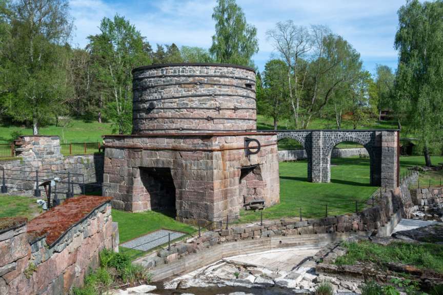 Eine historische Industrieanlage mit einem runden Steingebäude, Bögen und Mauern ist von Bäumen und grünem Gras umgeben.