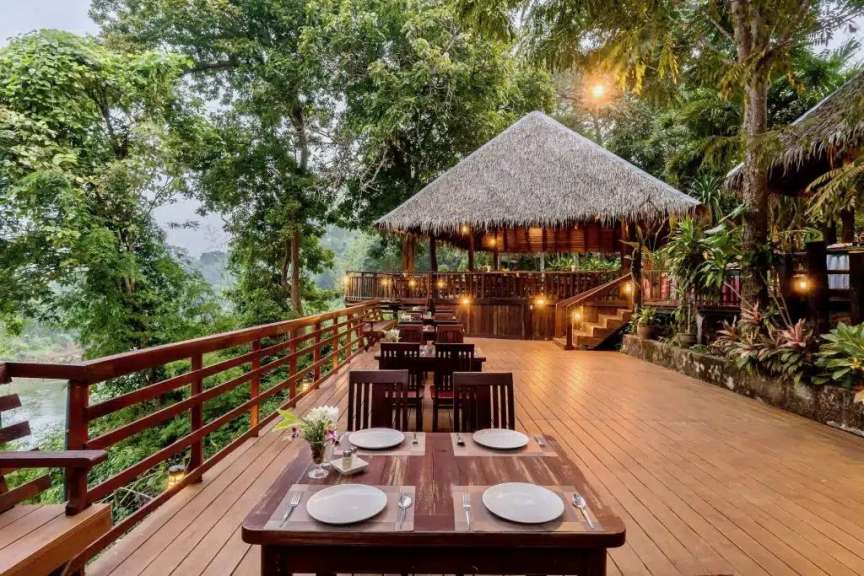 Ein Restaurant mit Strohdach und Holzdielen-Terrasse ist von üppiger Vegetation umgeben und blickt auf einen Fluss.