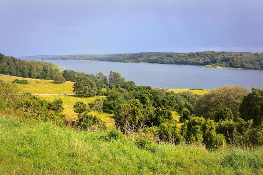 Ein breiter, ruhiger Fluss schlängelt sich durch eine hügelige, bewaldete Landschaft, mit grünen Wiesen und Bäumen im Vordergrund.
