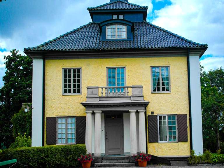 Eine gelbe Villa mit einem dunklen Dach und einem Balkon über dem Eingang wird von Bäumen und einem bewölkten Himmel umgeben.