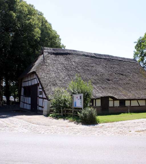 Ein langes, reetgedecktes Fachwerkhaus mit einem hohen Dach und einer Holzfassade steht unter einem blauen Himmel, daneben einige Bäume und ein Informationsschild.