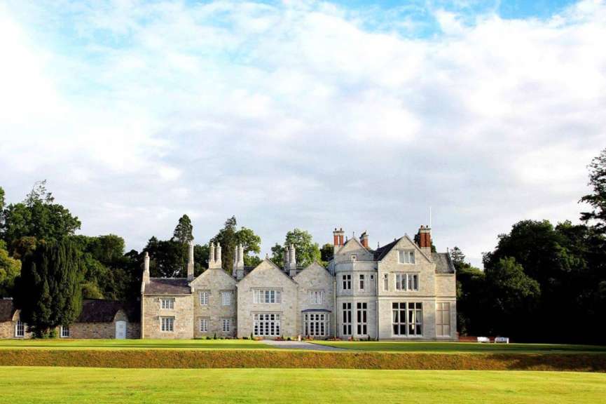 Ein großes, steinernes Herrenhaus mit vielen Fenstern und Schornsteinen steht vor einem grünen Rasen und Bäumen unter einem bewölkten Himmel.
