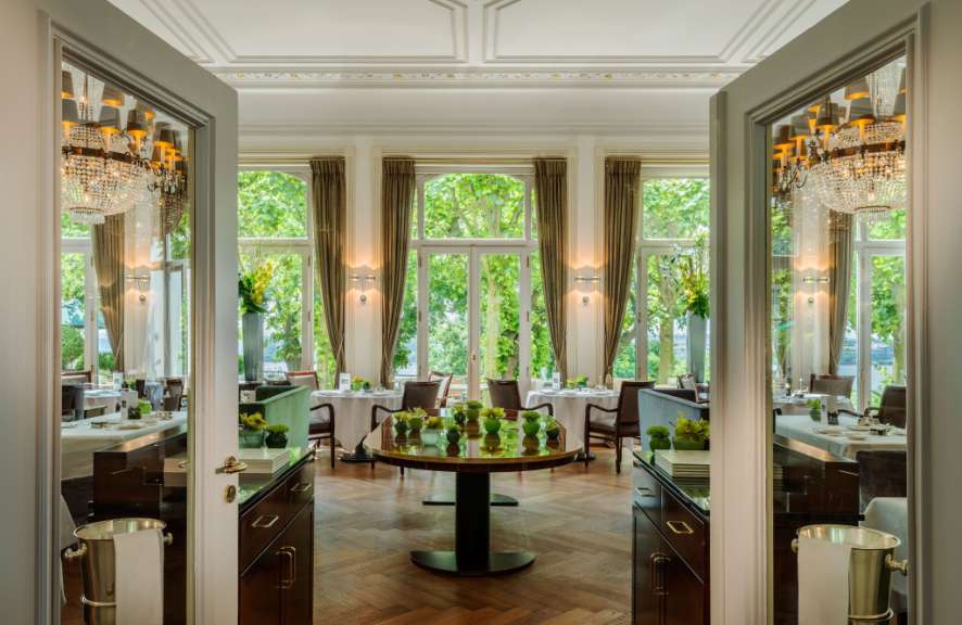 Eine elegante Speisesaal mit Spiegeln, einem runden Tisch und großen Fenstern mit Blick auf Bäume.