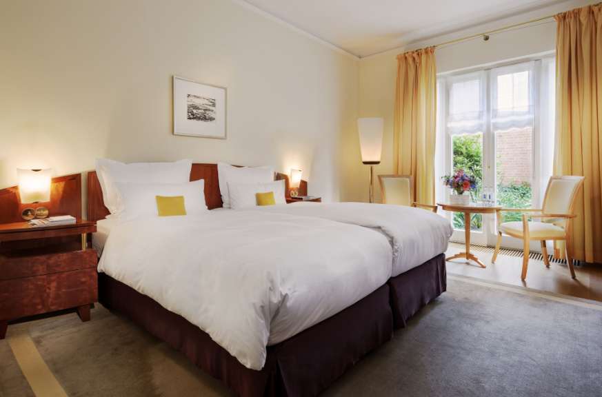 Ein Doppelbettzimmer mit zwei Betten, einem Nachttisch und einem Fenstersitzbereich wird in einem hellen, warmen Licht gezeigt.
