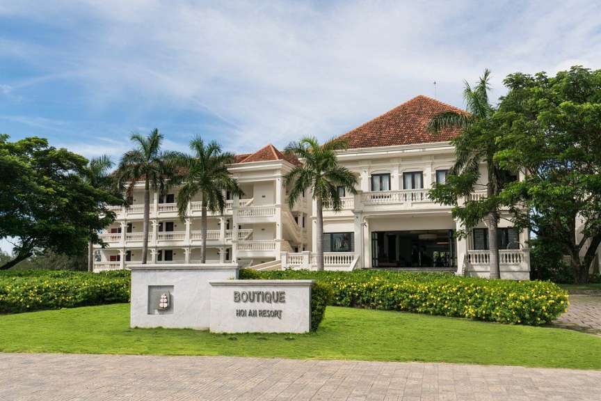 Ein großes weißes Hotel mit roten Ziegeldächern und Palmen steht vor einem blauen Himmel mit Wolken.