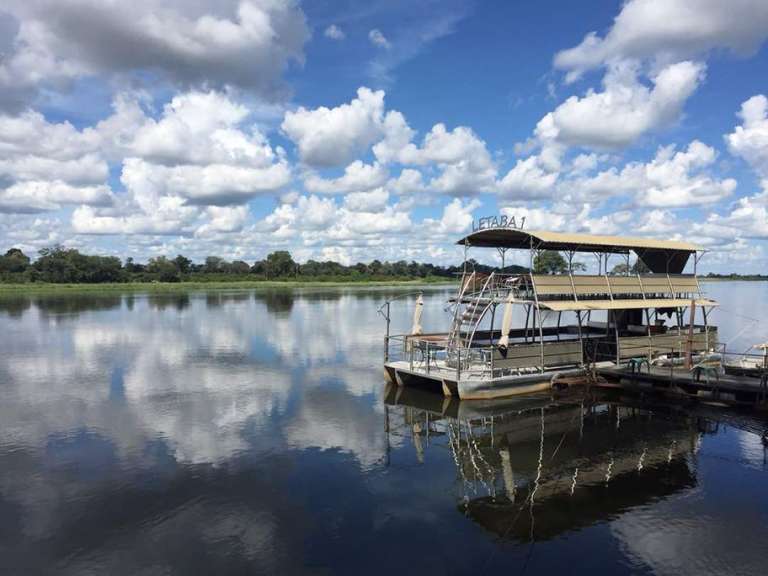 Ein zweistöckiges Boot namens LETABA 1 liegt am Kai an einem Fluss vor einer grünen Baumkulisse unter einem blauen Himmel mit Wolken.