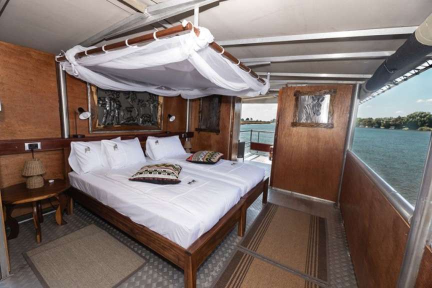 Ein Schlafzimmer auf einem Boot mit zwei Betten, einem Baldachin, zwei Kissen und einer Aussicht auf das Wasser.