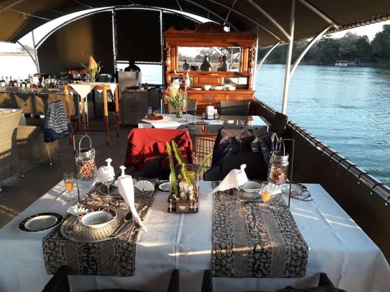 Ein gedeckter Tisch auf einem Boot mit Blick auf einen Fluss und eine bewaldete Uferlinie.