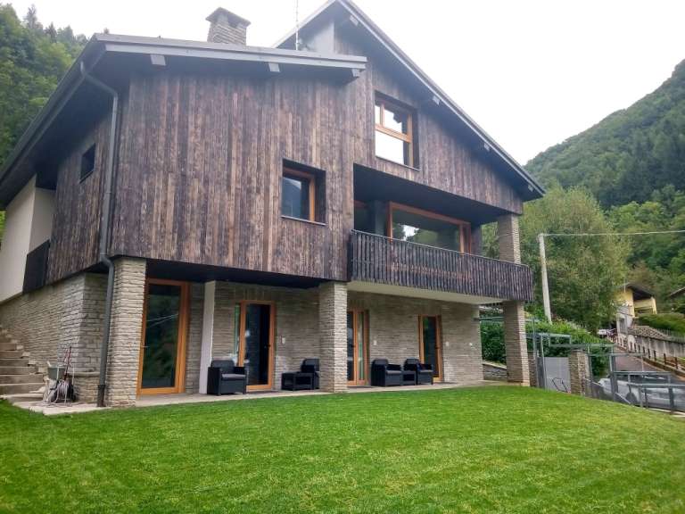 Ein modernes Holzhaus mit Steinfundament und einem Balkon ist von einem grünen Rasen und bewaldeten Hügeln umgeben.