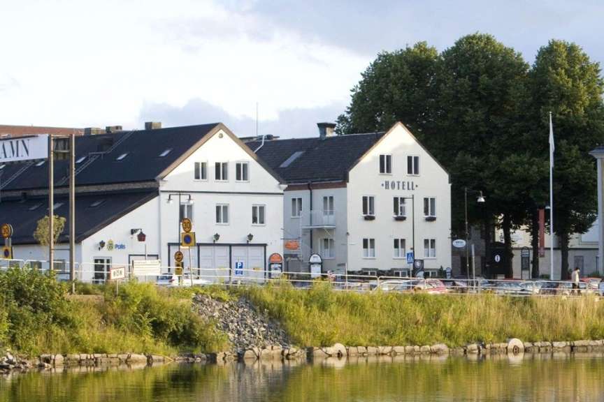 Ein Flussufer mit einer Uferbefestigung, bewachsenem Ufer und Gebäuden, darunter ein Hotel, erstreckt sich im Hintergrund.