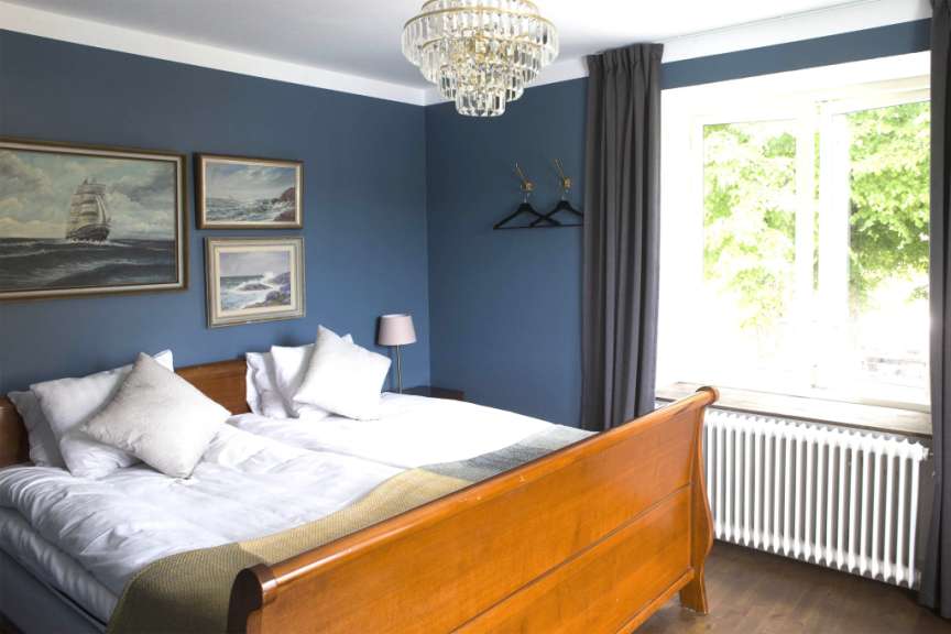 Ein luxuriöses Schlafzimmer mit blau gestrichenen Wänden, einem großen Bett mit weißen Laken und Kissen, einem Holzbettrahmen, einem Kronleuchter, drei Gemälden von Segelschiffen und einer stehenden Lampe.