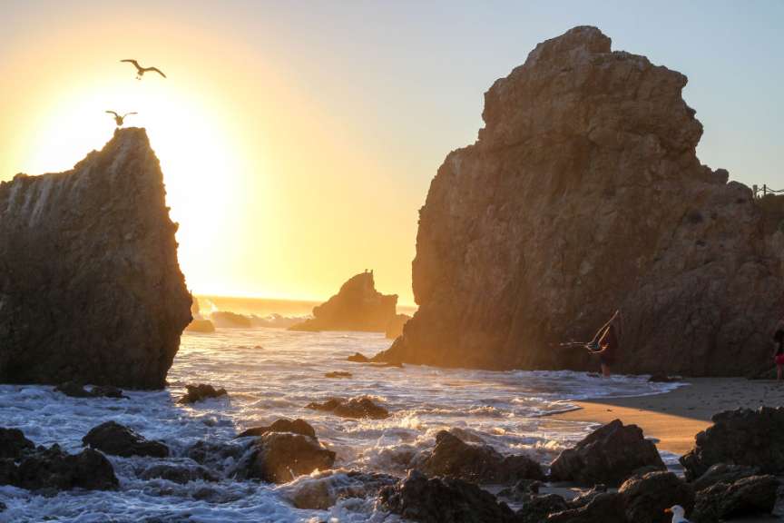 Sonnenlicht beleuchtet einen Strand mit Felsen, Möwen und aufziehenden Wellen.