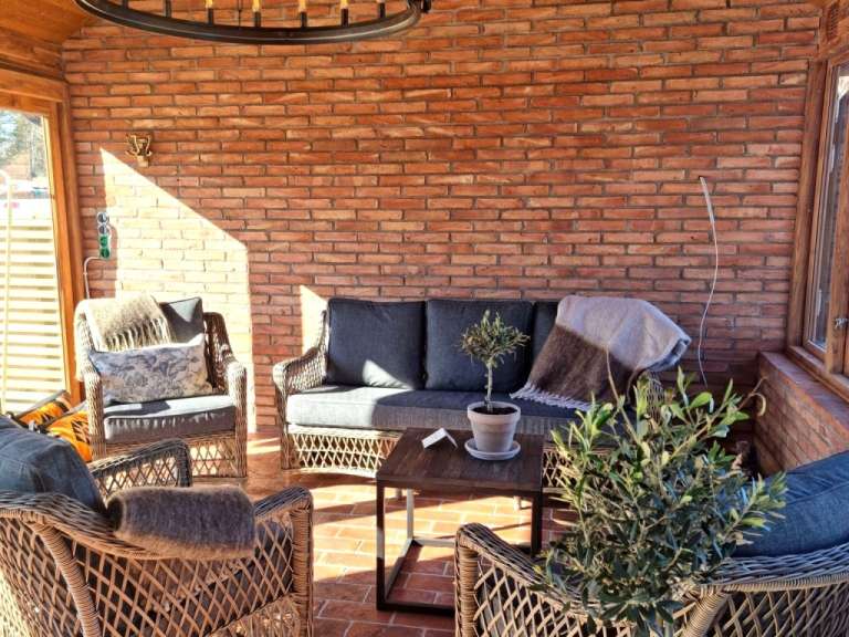 Auf der überdachten Veranda stehen Rattansessel und ein Sofa vor einer Ziegelwand, beleuchtet von Sonnenlicht, das durch die Jalousien fällt.