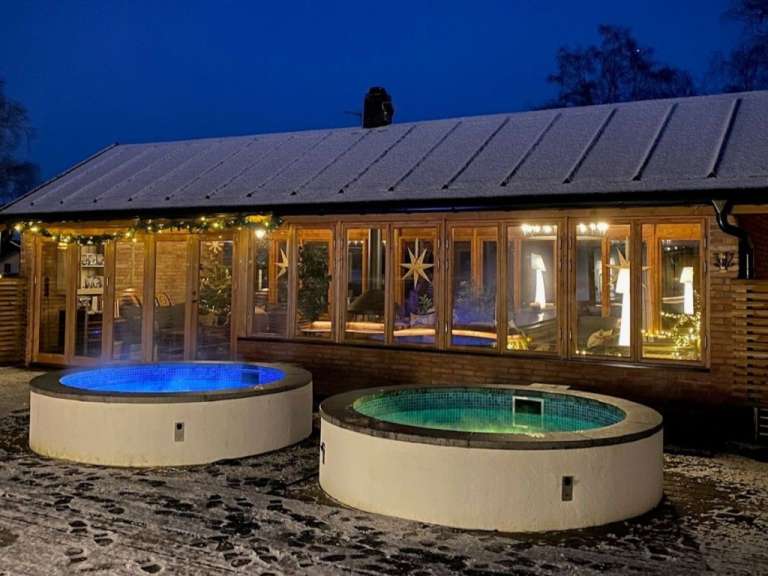 Zwei beleuchtete Whirlpools stehen im Schnee vor einem beleuchteten Haus mit großen Fenstern, das im Dunkeln mit Weihnachtslichtern geschmückt ist.